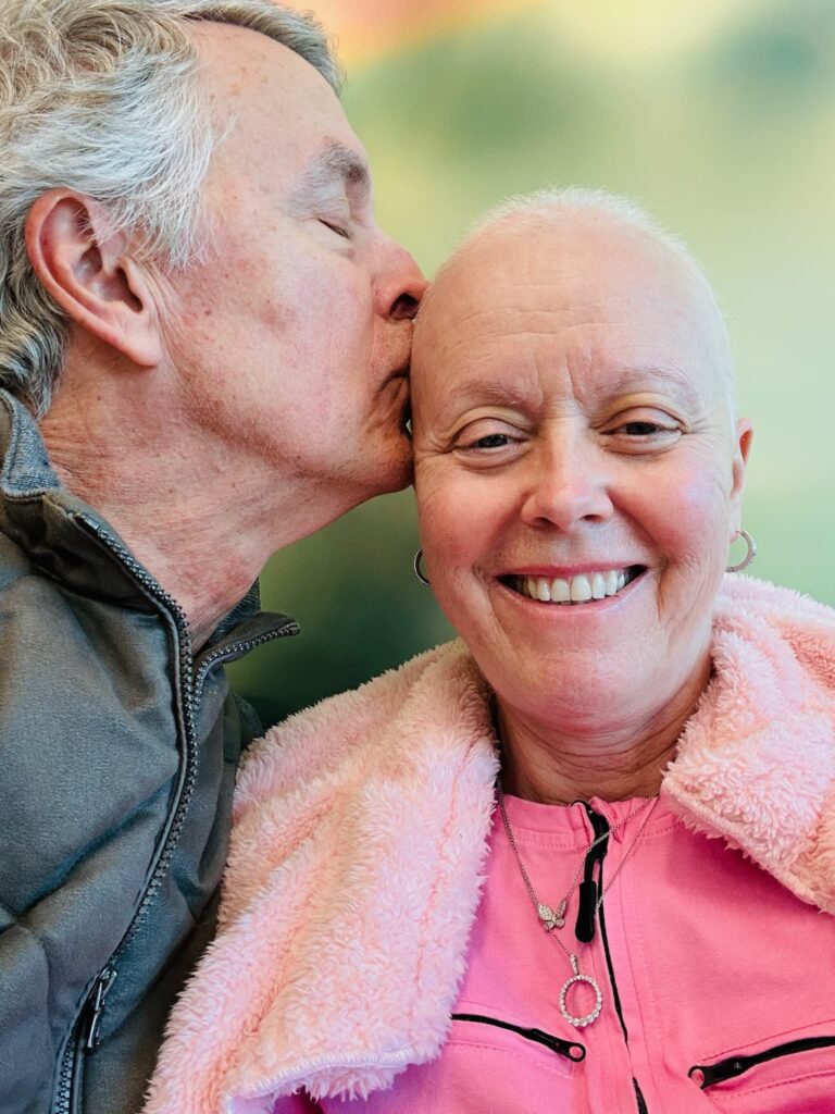 Elderly man kisses smiling bald woman on head.